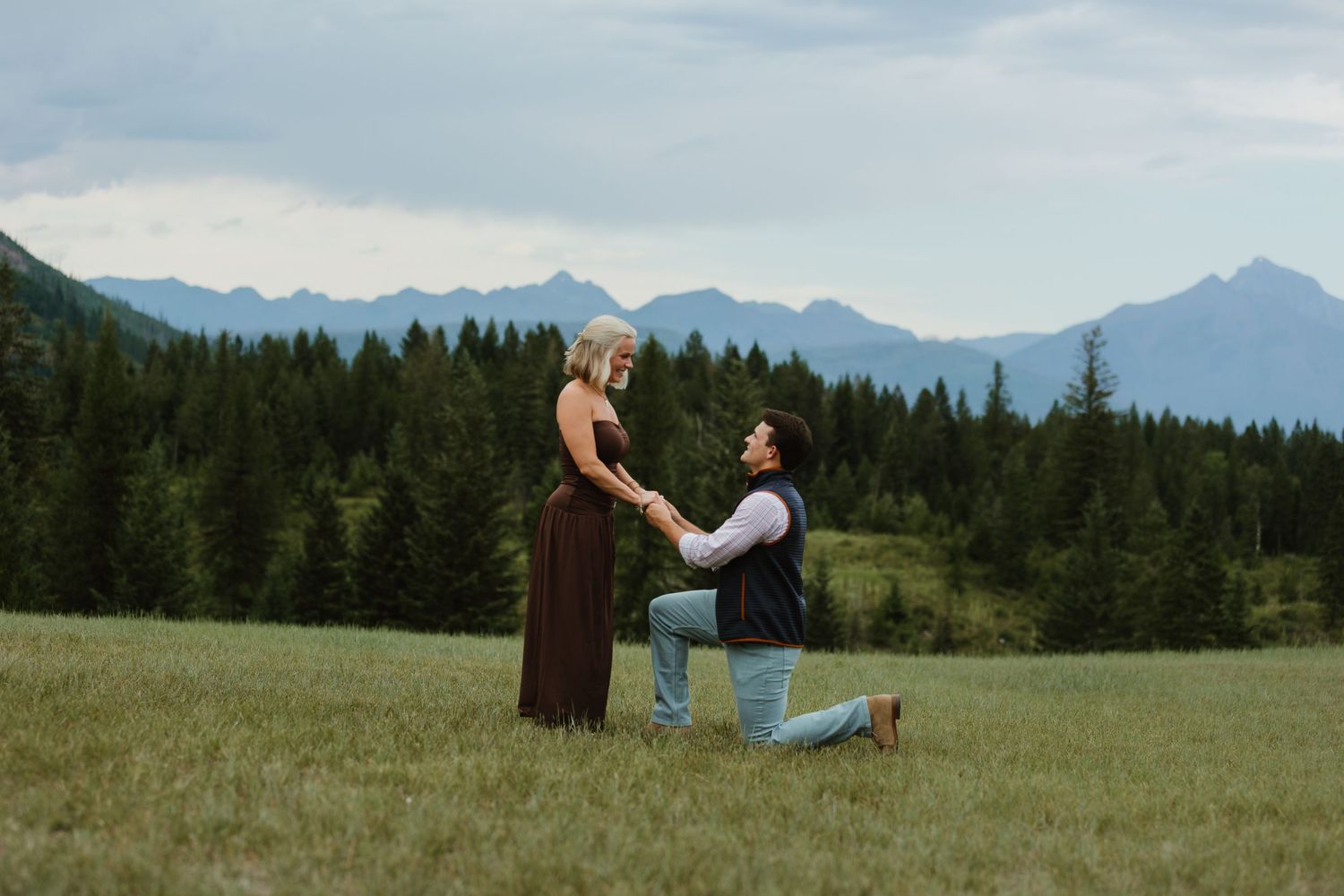 Man proposes on one knee to woman in mountain meadow with pine trees and Rocky Mountains in background.