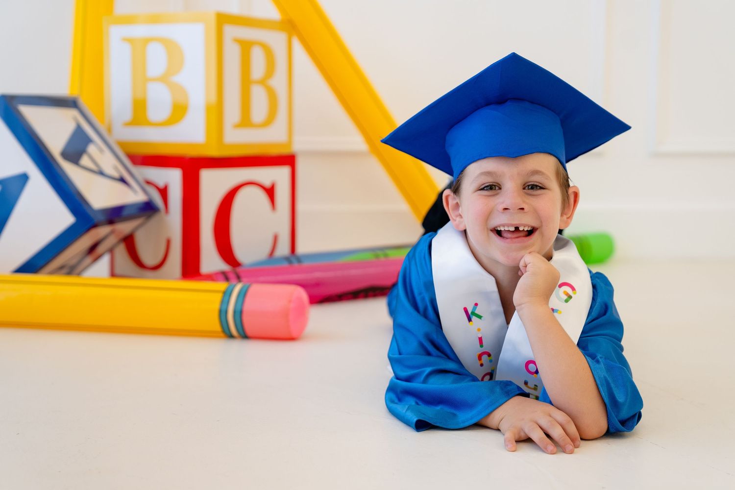 Young child in blue graduation cap and gown smiling beside alphabet blocks and school supplies.