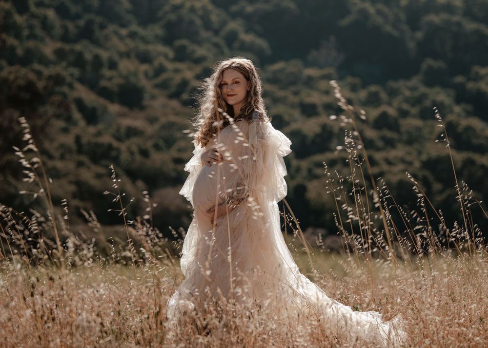 Woman in white lace maternity gown standing in golden field with lush green hills in background.