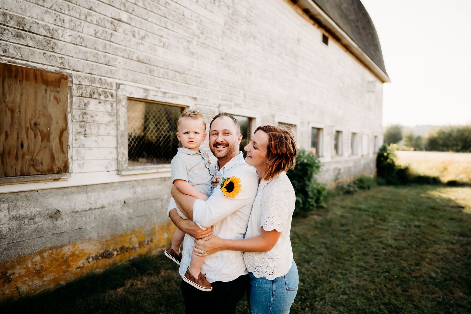 Happy family with baby poses near rustic barn holding sunflowers on a sunny day. Family photos in Tacoma, WA.