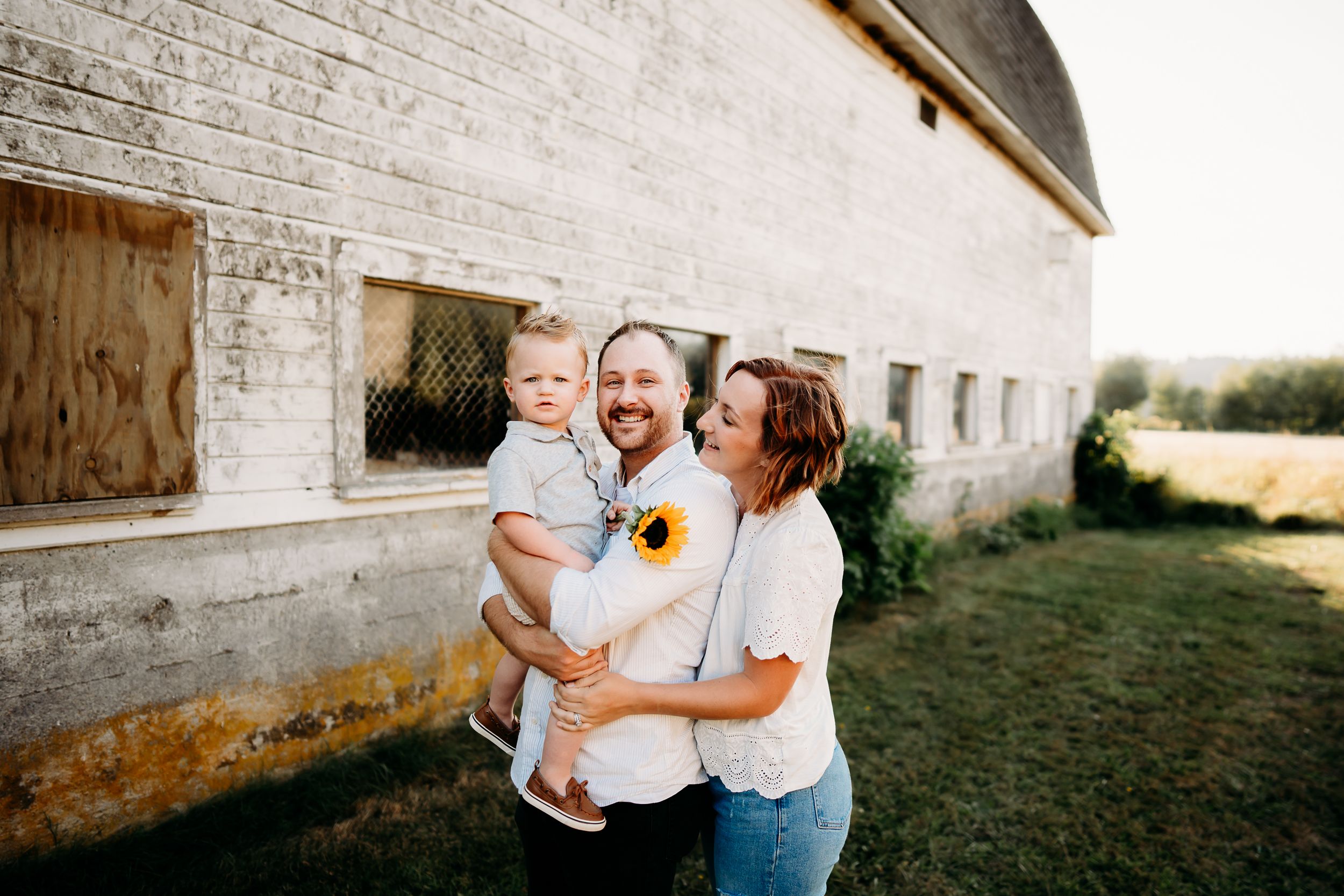 Happy family with baby poses near rustic barn holding sunflowers on a sunny day. Family photos in Tacoma, WA.