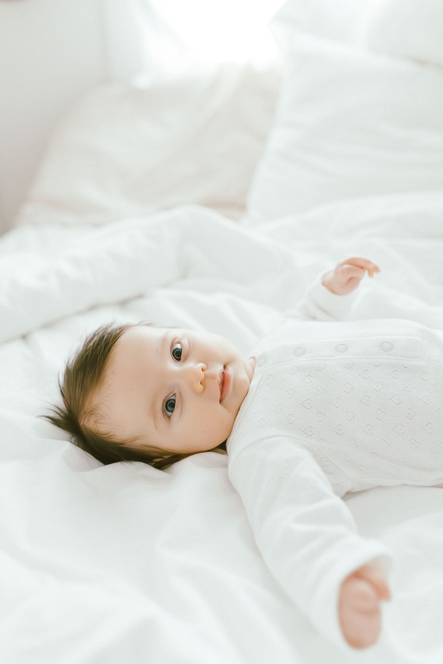 A cute baby in a white onesie lying on white bedding, looking up with bright curious eyes.
