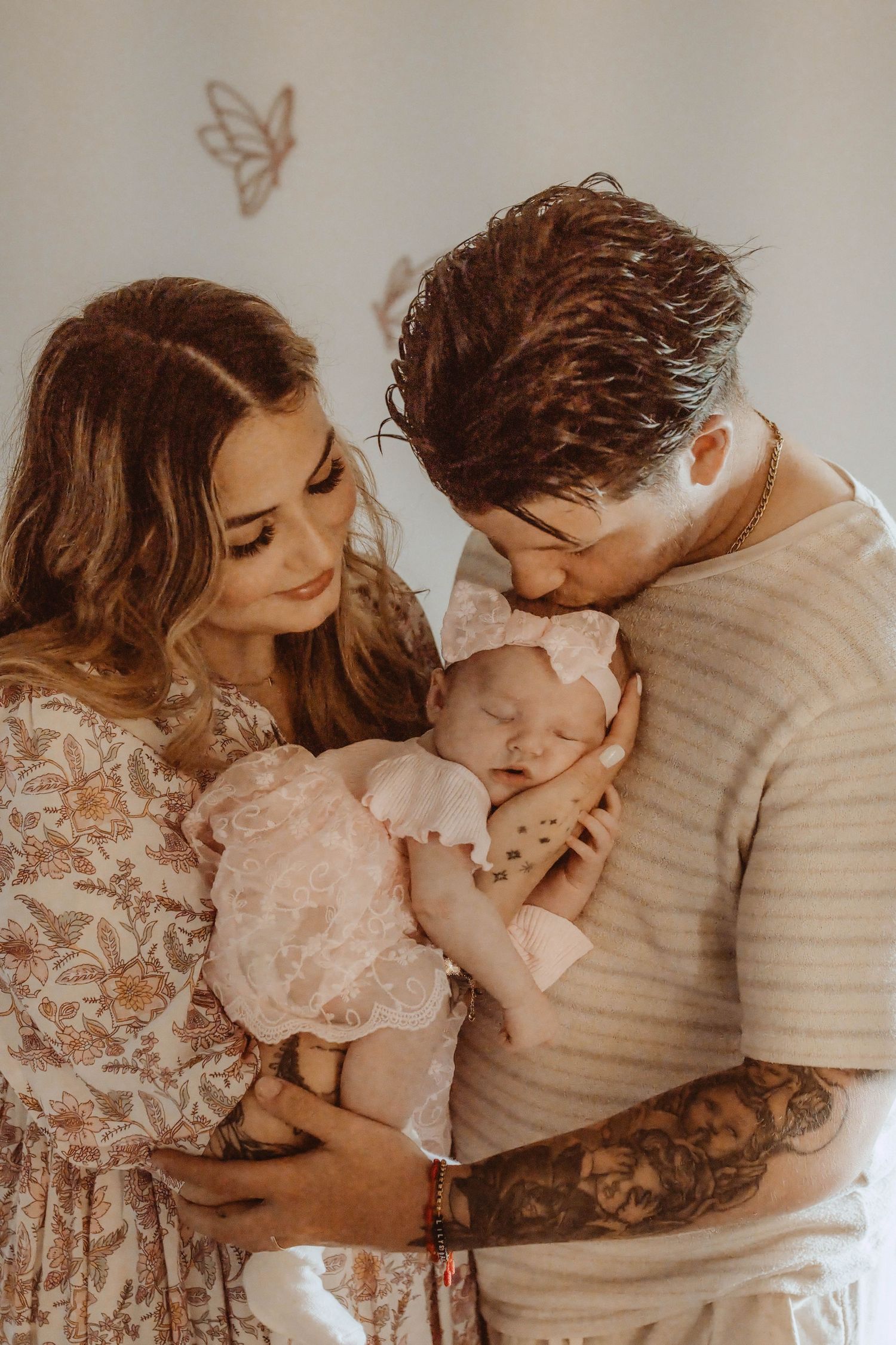 New parents tenderly hold their sleeping newborn baby girl in pink lace outfit, father kissing her head.
