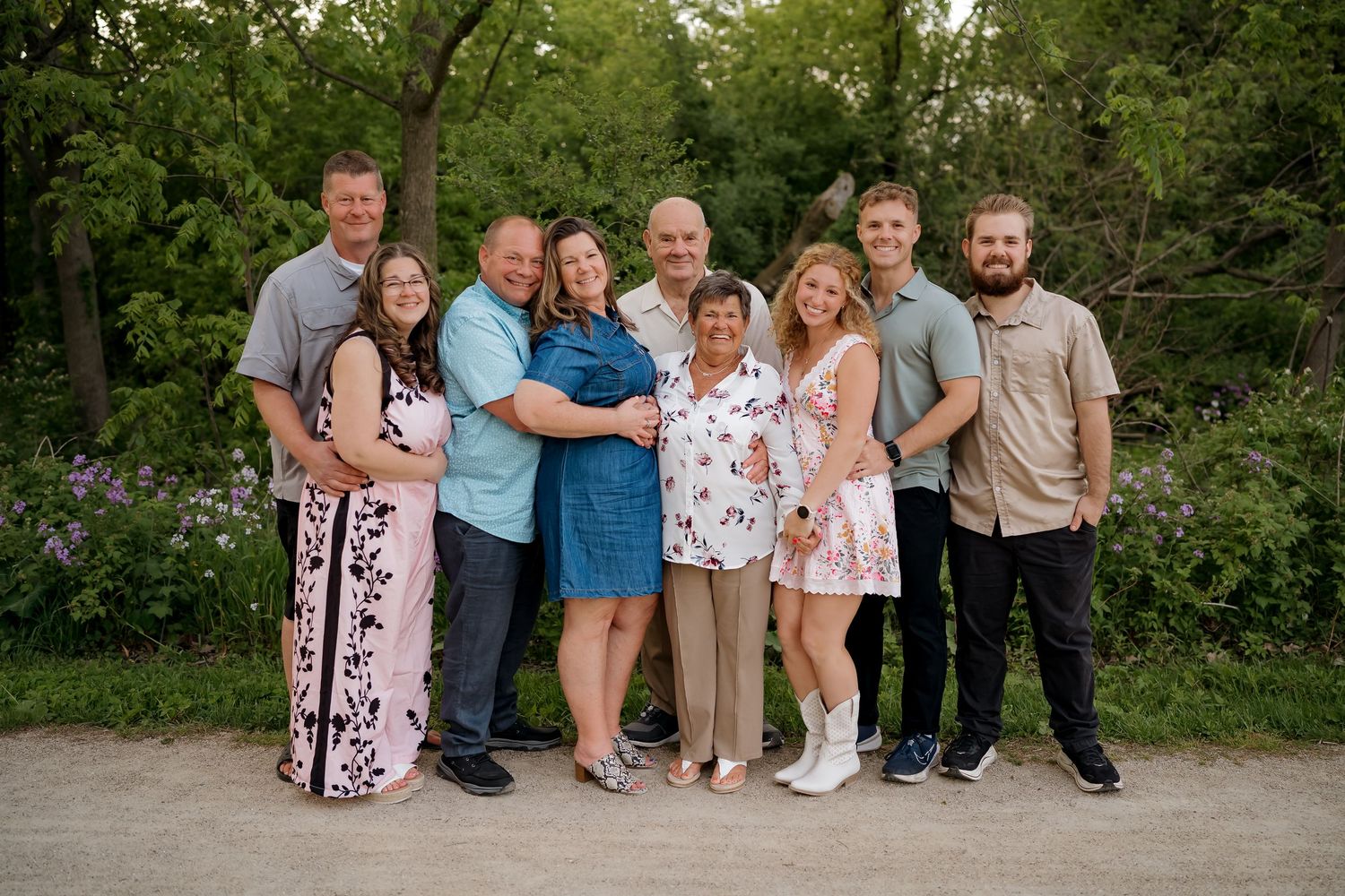 A large family of nine poses together outdoors on a path surrounded by lush green trees and purple wildflowers.