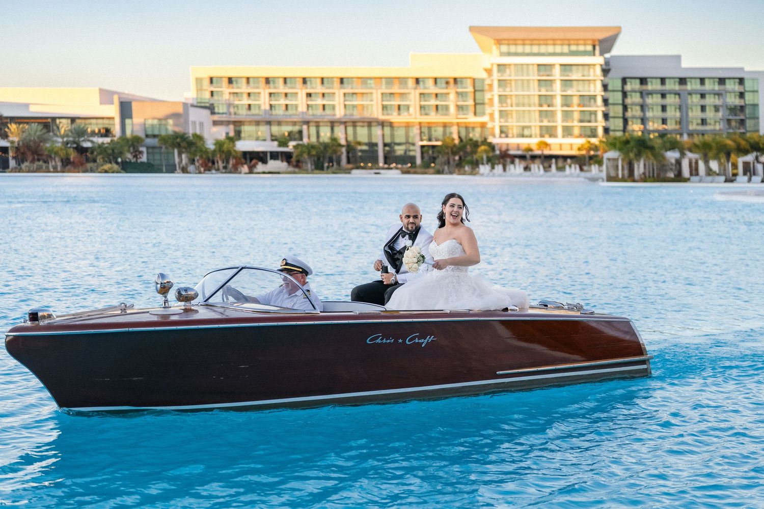 Bride and groom riding a classic wooden Chris-Craft boat on blue water with a luxury resort in the background.