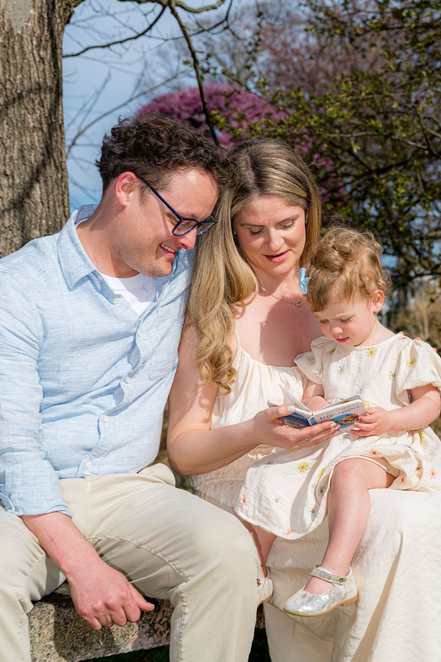 A happy family of three sits outdoors, reading a book together near a blooming tree on a sunny day.