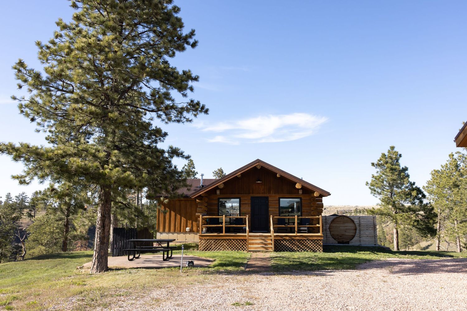 Rustic log cabin with wrap-around porch surrounded by pine trees under a clear blue sky on a gravel lot.
