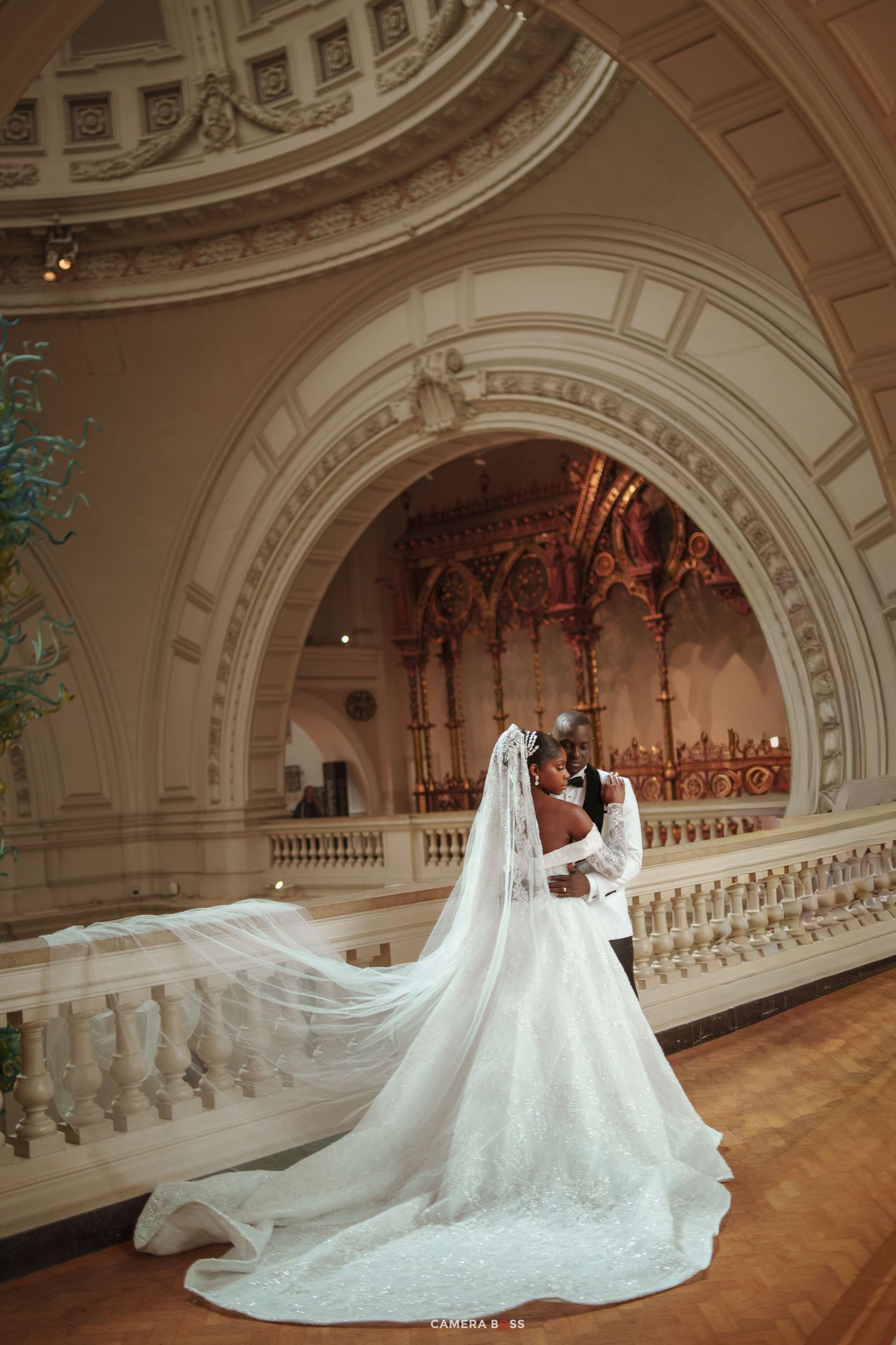 Bride and groom share a romantic kiss in an ornate ballroom, her long lace veil trailing elegantly across the marble floor.