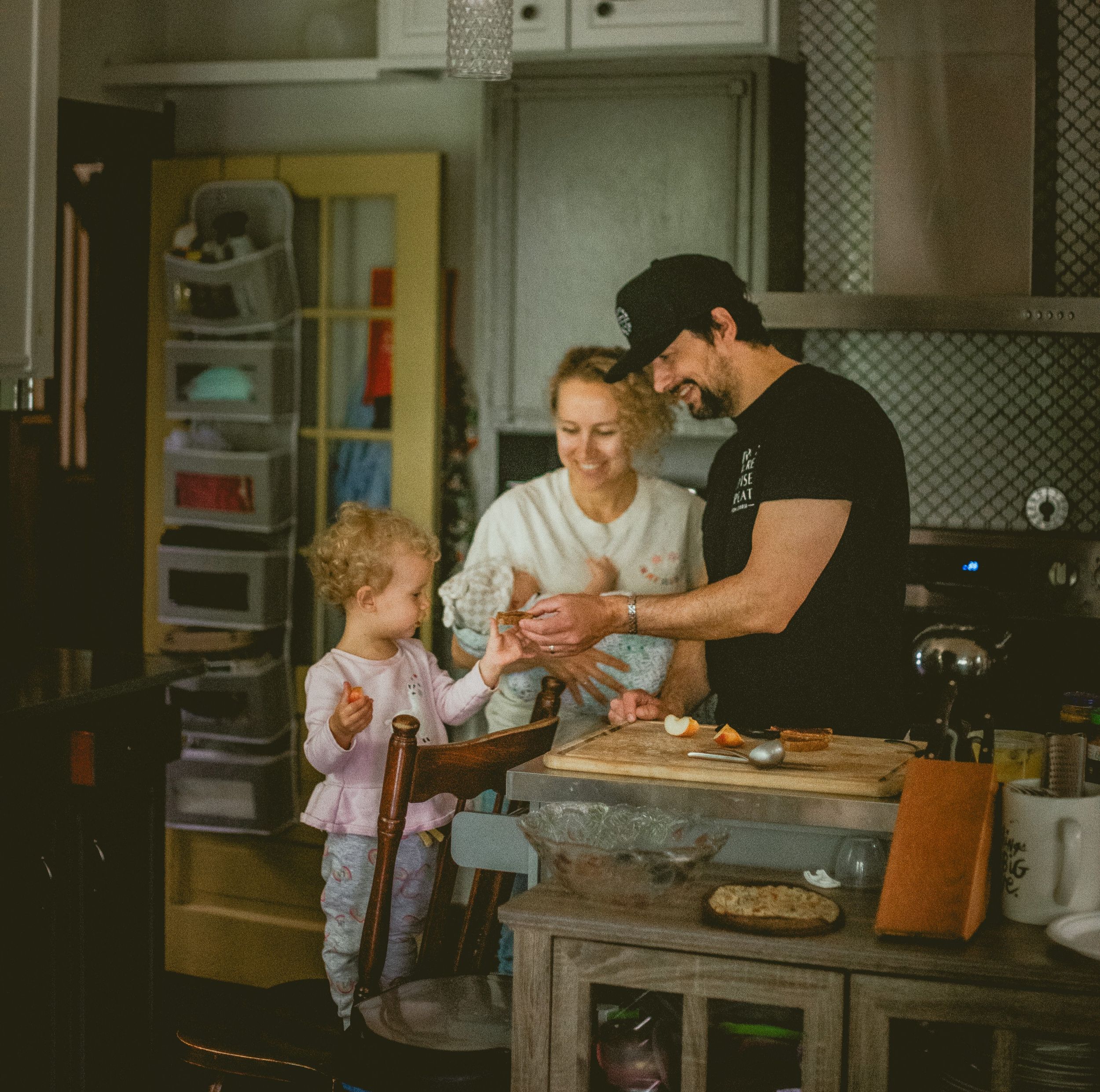 A couple shares a tender kiss while holding a baby in a rustic kitchen, with a young child playing nearby. In-home Documentary Family Photos