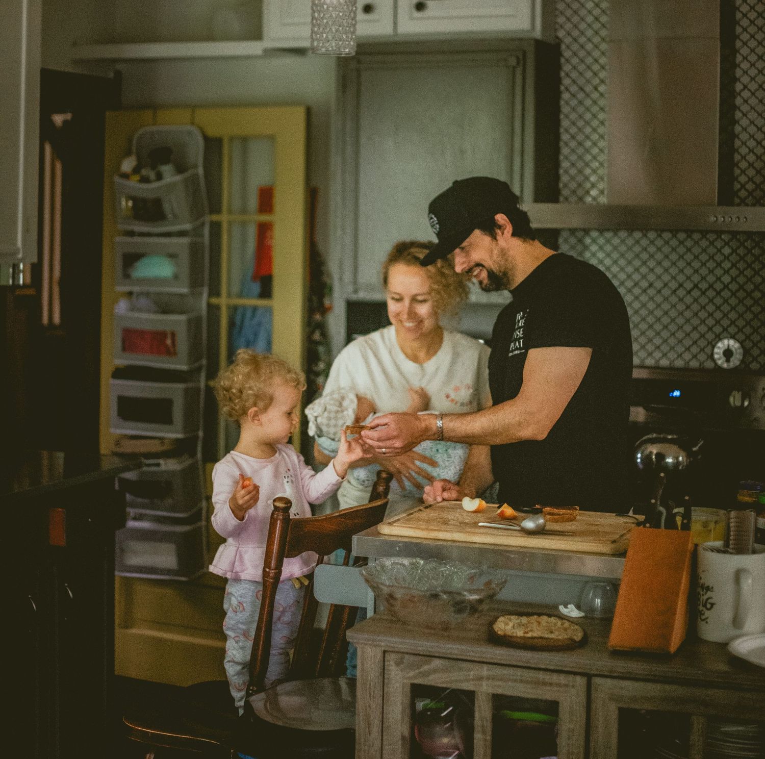 A couple shares a tender kiss while holding a baby in a rustic kitchen, with a young child playing nearby. In-home Documentary Family Photos