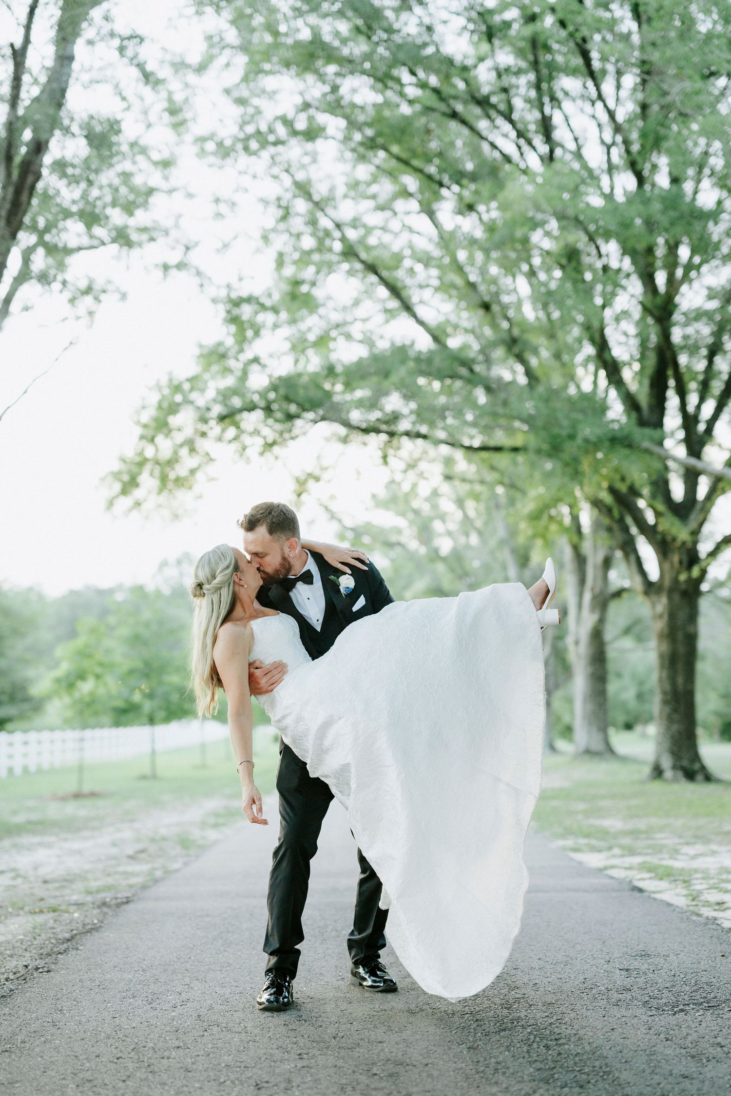 Groom in black tuxedo lifts bride in white gown while kissing on tree-lined path at outdoor wedding.