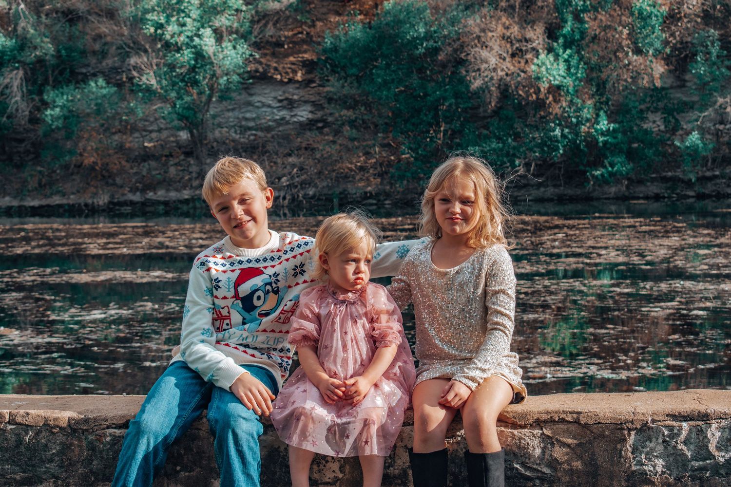 Three children sitting on a stone wall outdoors, with a boy in a Christmas sweater and two girls in dresses.