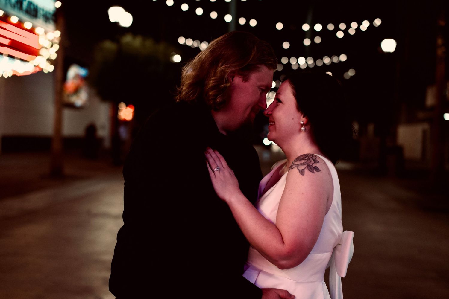 Elegant couple sitting on steps under sparkling lights, woman in white dress and heels, man in black vest, romantic ambiance.