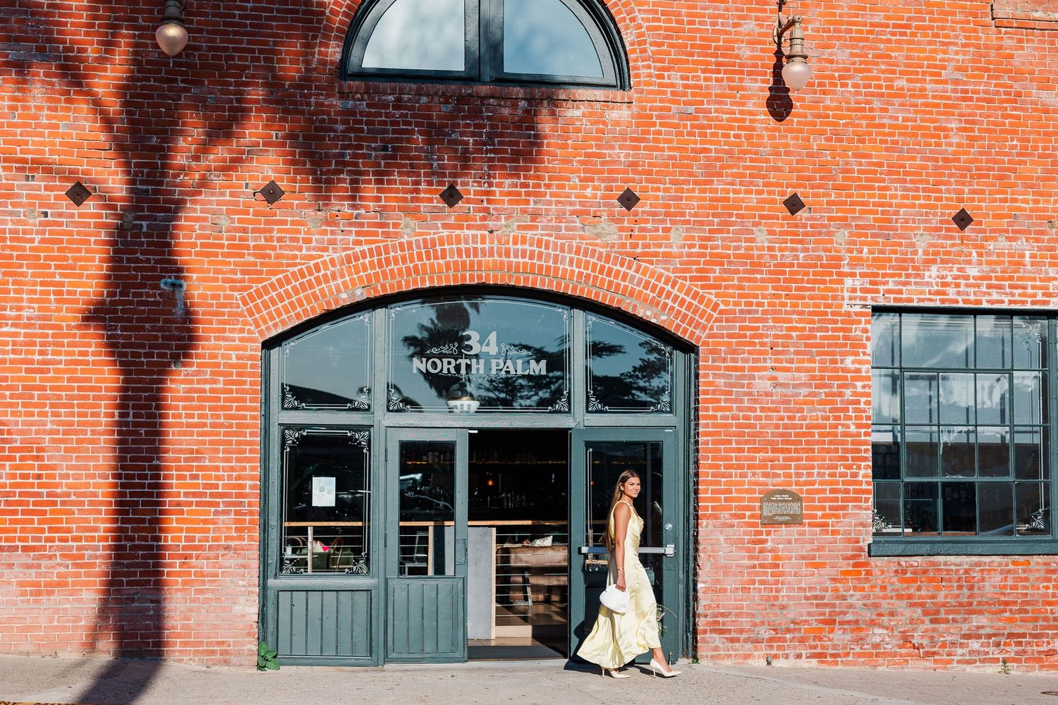 Woman walking past a red brick building with gray doors at North Elm restaurant entrance.