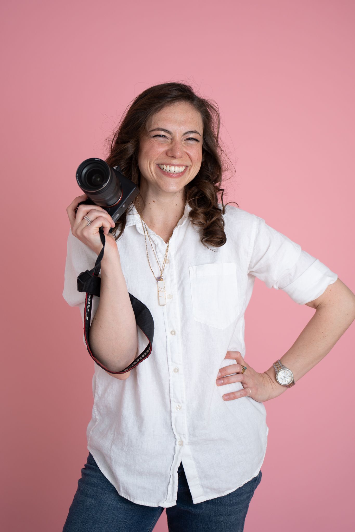 Smiling woman in white shirt holding a DSLR camera on pink background, perfect for photography business branding.