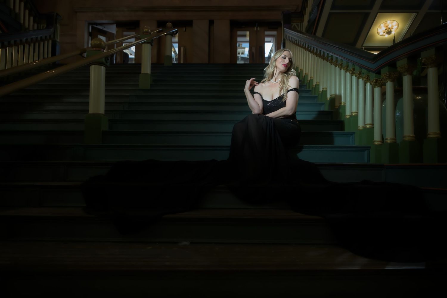 Elegant woman in black gown seated on dimly lit grand staircase, dramatic lighting creating moody atmosphere.