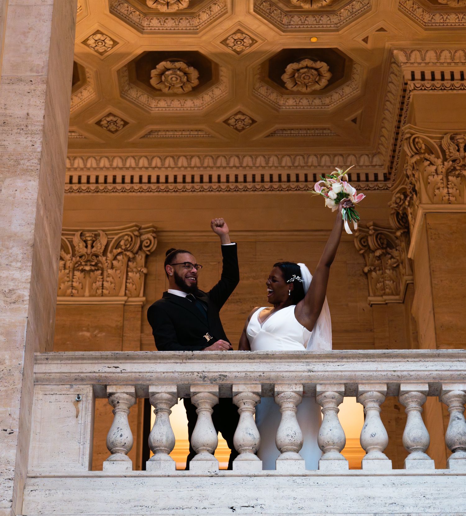 Joyful newlyweds celebrate on ornate marble balcony, bride holding bouquet aloft in elegant wedding venue.