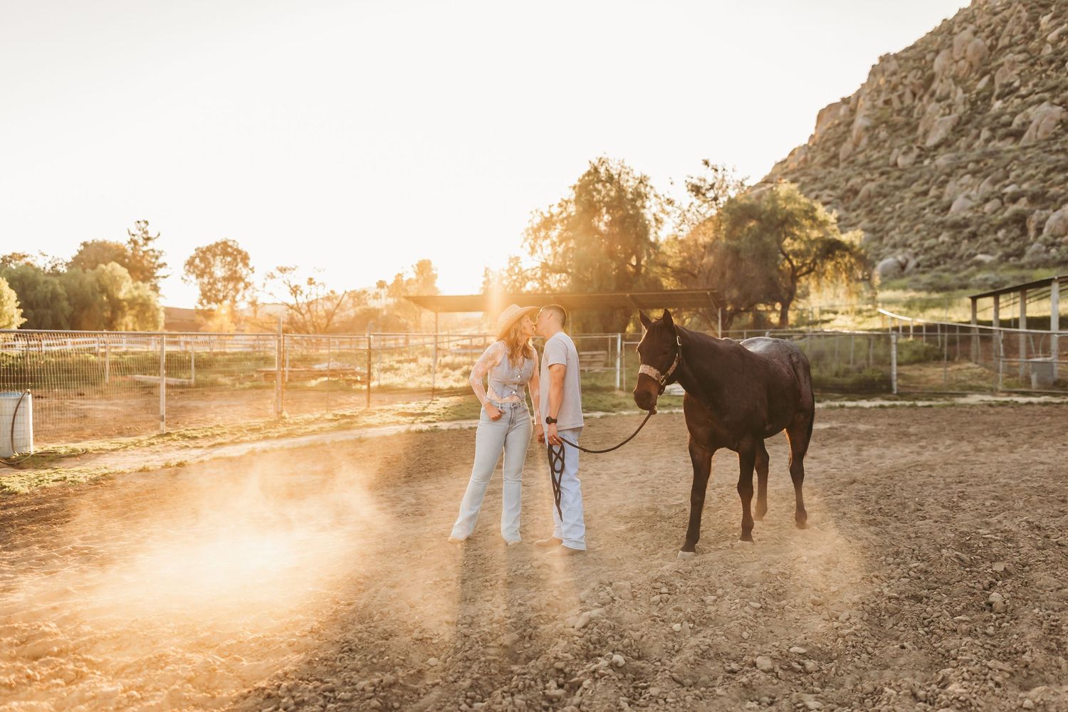 Two people stand with a dark horse at golden hour on a dusty ranch with rocky hills in the background.