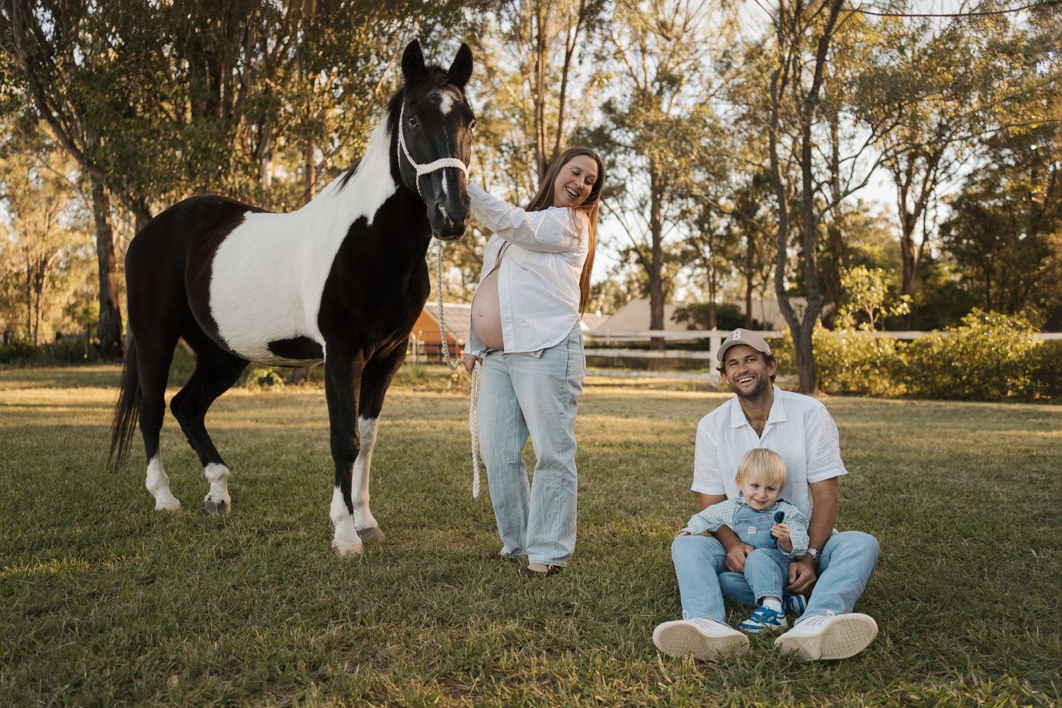 Family with toddler poses beside a black and white paint horse on green grass surrounded by trees during a maternity photoshoot in the hills
