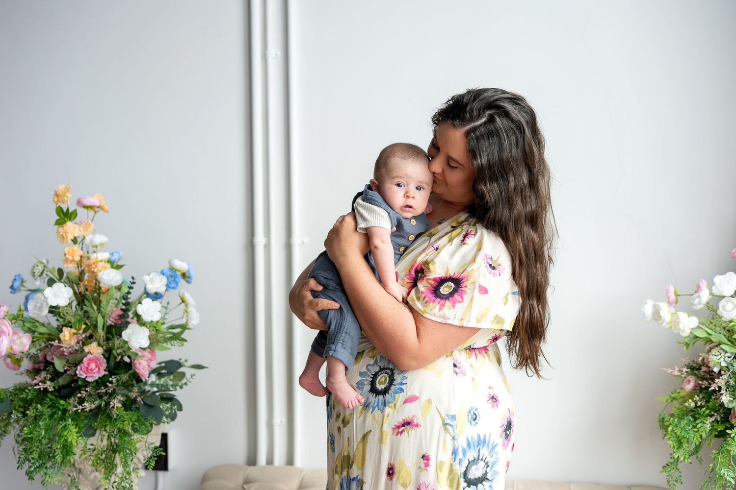 Loving mother in floral dress kissing her newborn baby in bright studio with spring flowers.