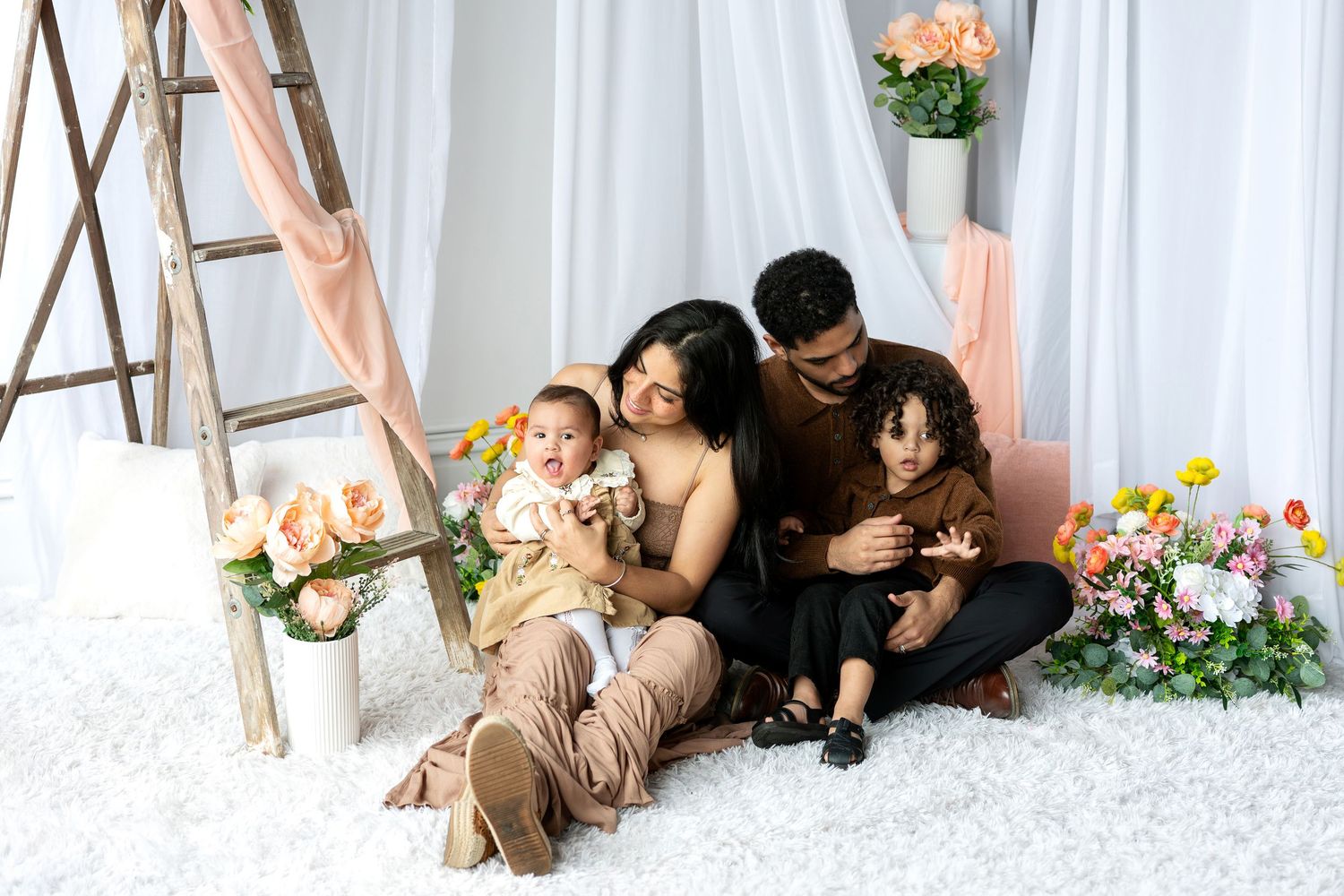 Family of four with two young children sit together on white rug with flowers and ladder.