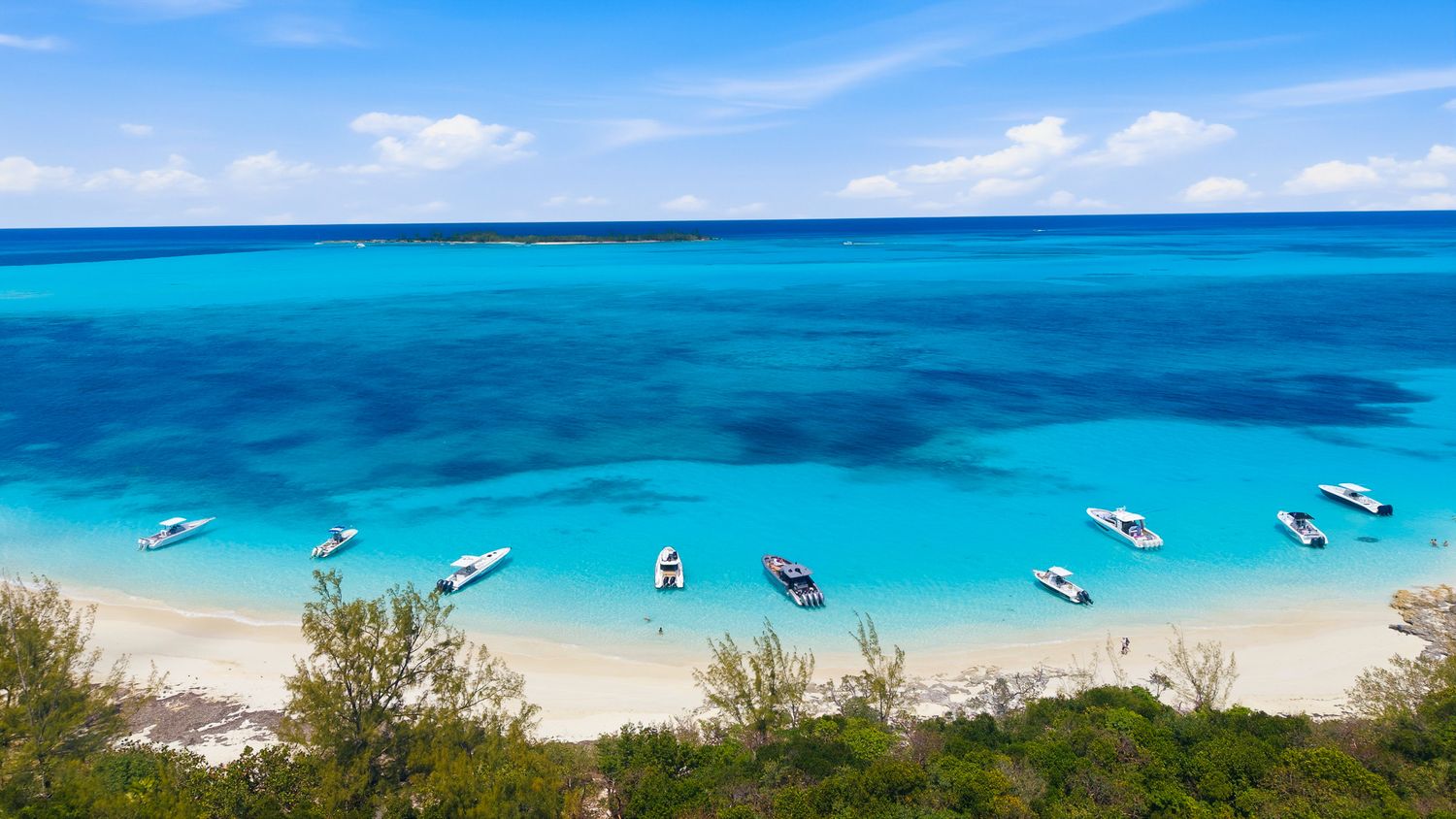 Aerial view of a pristine tropical beach with turquoise waters and white sand, dotted with small boats under a blue sky.