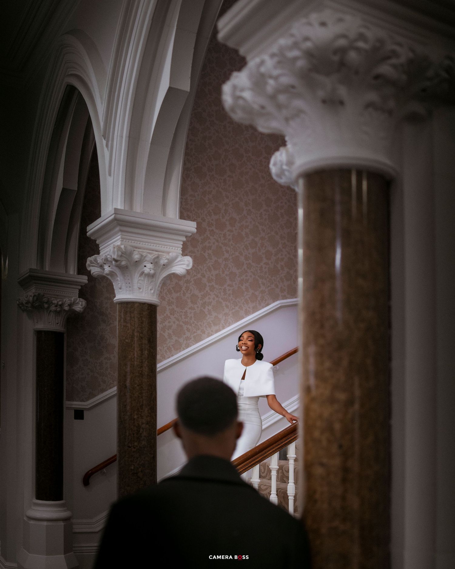 Bride in white dress descends grand staircase with ornate columns in elegant historic venue.