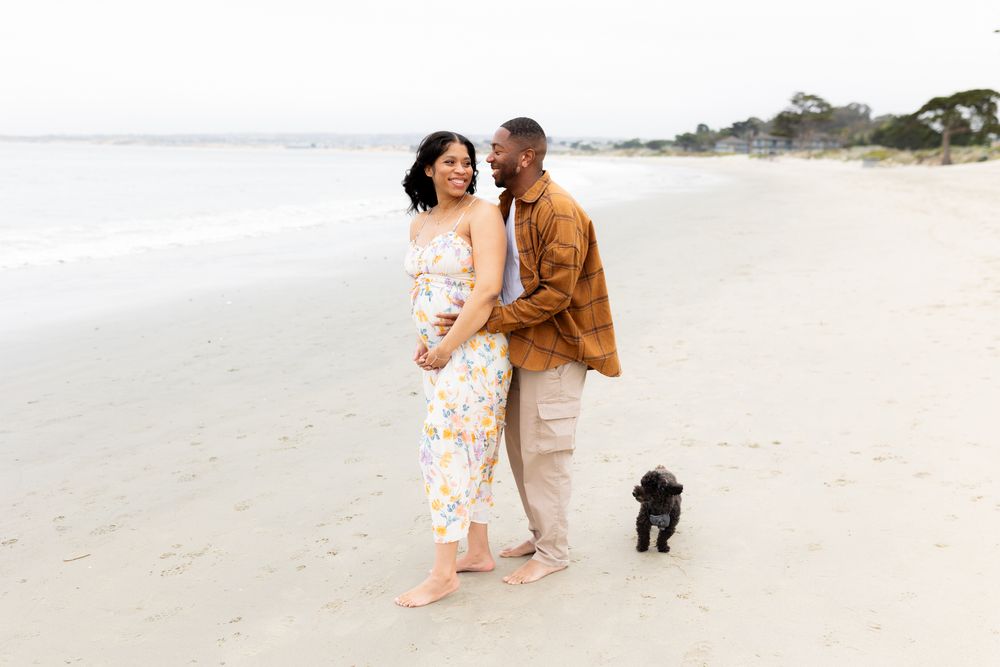 A couple walks barefoot on a sandy beach with a small black dog, the woman in a floral jumpsuit, man in brown shirt.