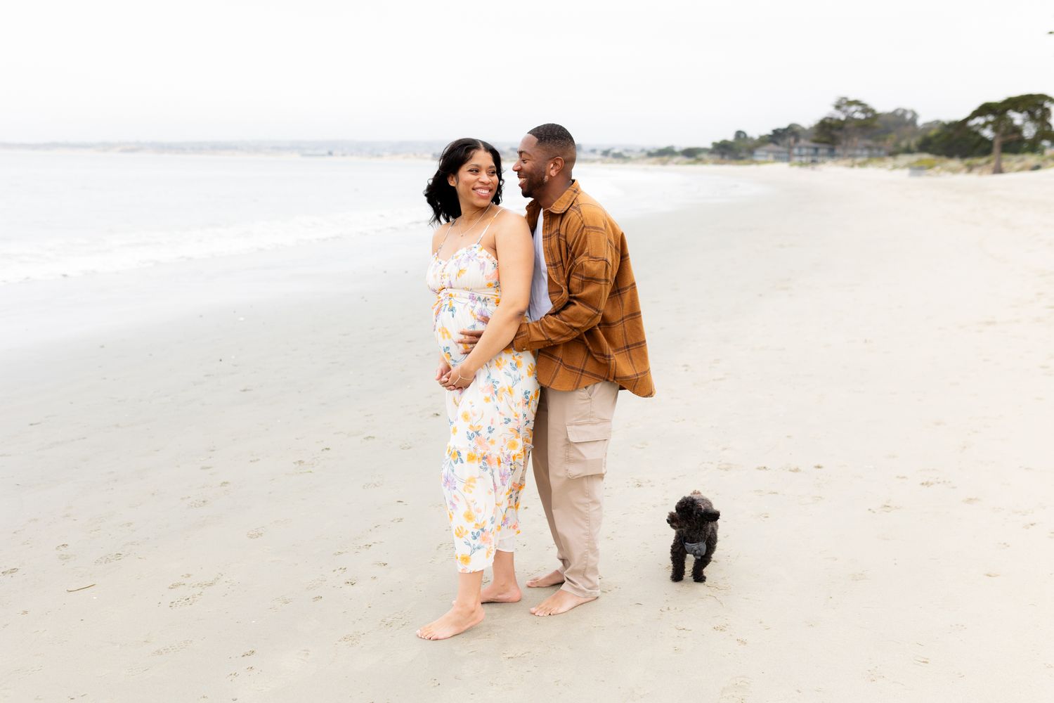 A couple walks barefoot on a sandy beach with a small black dog, the woman in a floral jumpsuit, man in brown shirt.