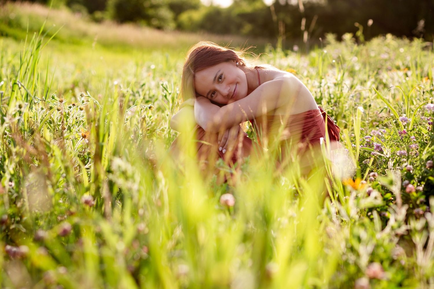Young woman with red hair relaxing in a sunny wildflower meadow, smiling peacefully among green grass and blooms.
