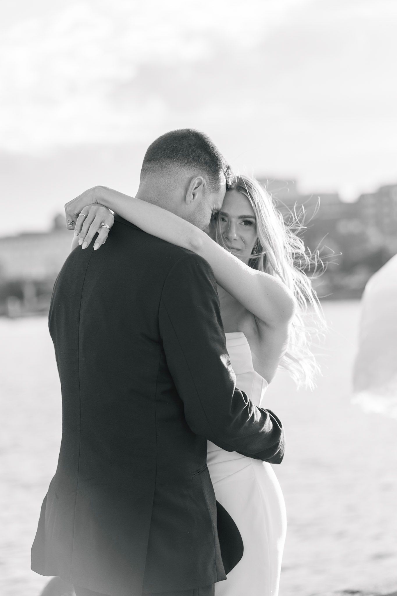 Black and white photo of newlyweds embracing on beach with bride smiling over shoulder.