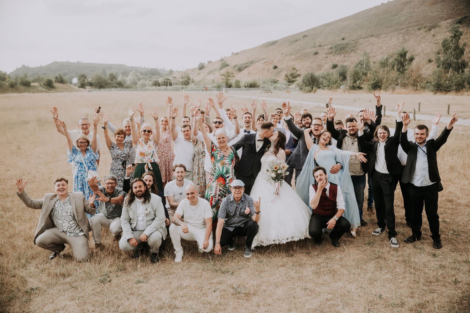 Wedding party celebrating outdoors in a field with rolling hills, bride and groom surrounded by cheering guests raising arms.