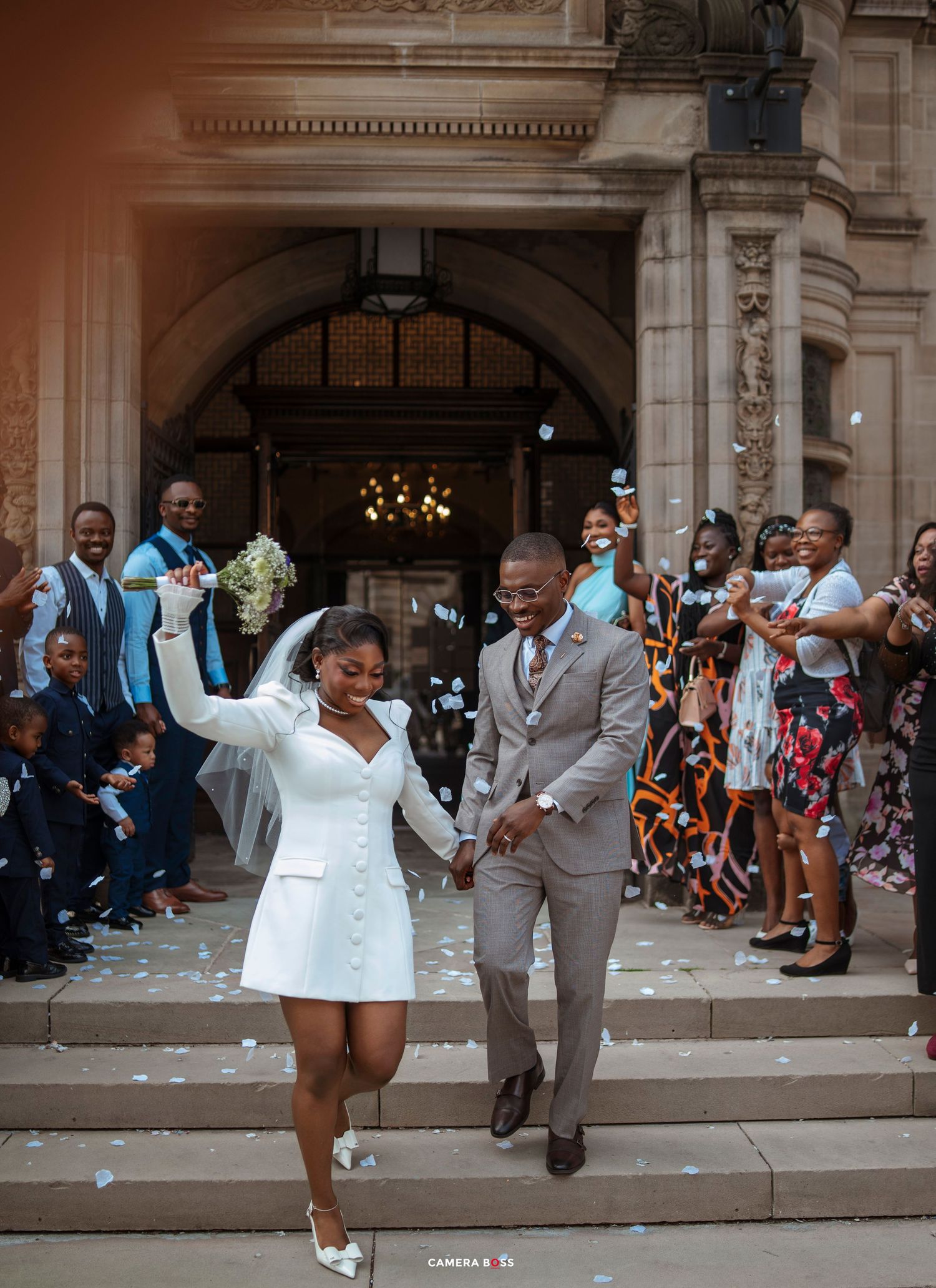 Joyful newlyweds exit grand building amid confetti shower, bride in white mini dress, groom in tan suit, guests celebrating.