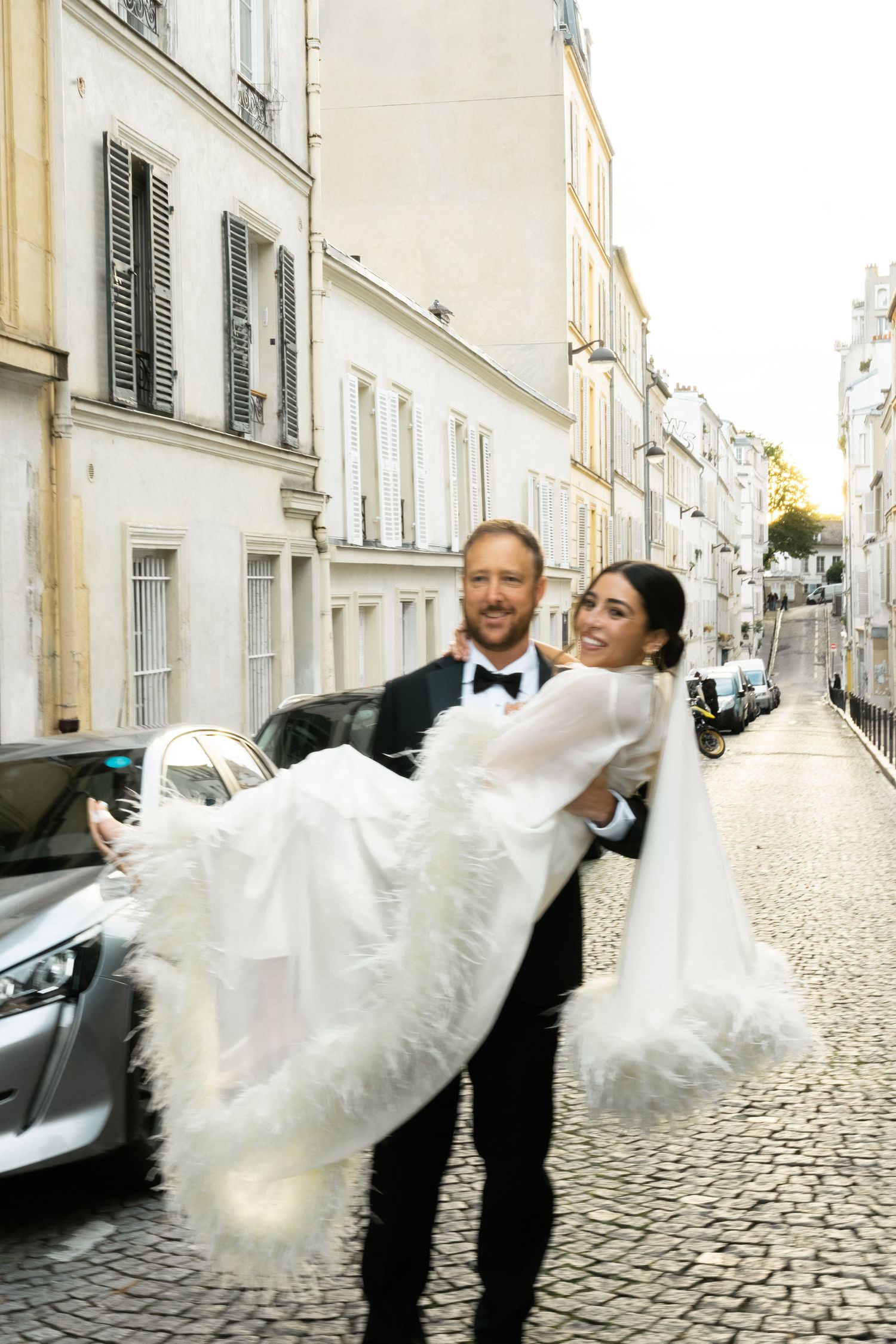 Groom carries bride in white feathered gown on cobblestone Paris street, both smiling joyfully on their wedding day.
