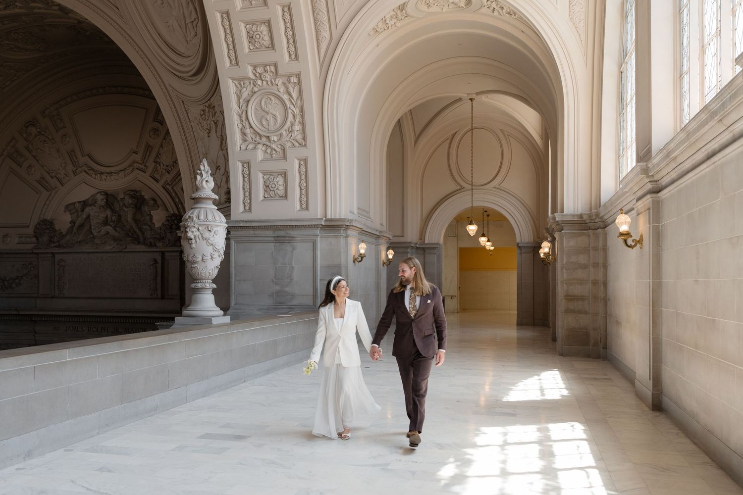 Bride and groom walking hand in hand through an elegant marble hallway at San Francisco City Hall wedding.