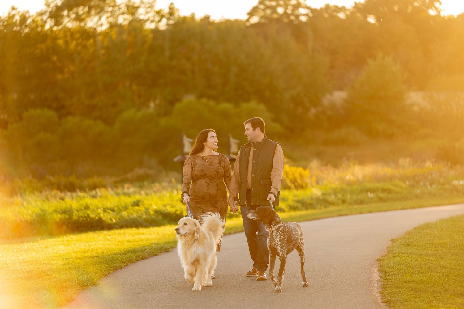 Couple walking two dogs on a paved path at golden hour sunset in a lush green park.