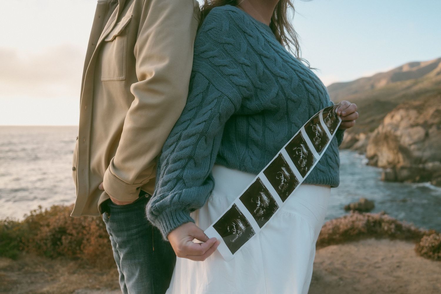 Couple holding hands on coastal cliffside with wildflowers at sunset, woman in white skirt and gray sweater, man in beige jacket.