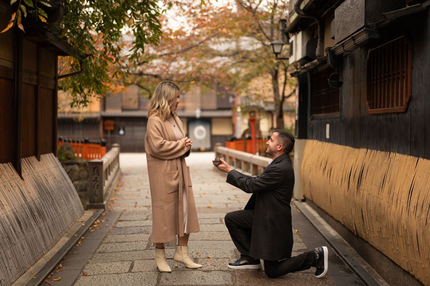 Man kneels on one knee proposing to a woman in a beige coat on a quiet stone-paved street lined with traditional wooden buildings in autumn.