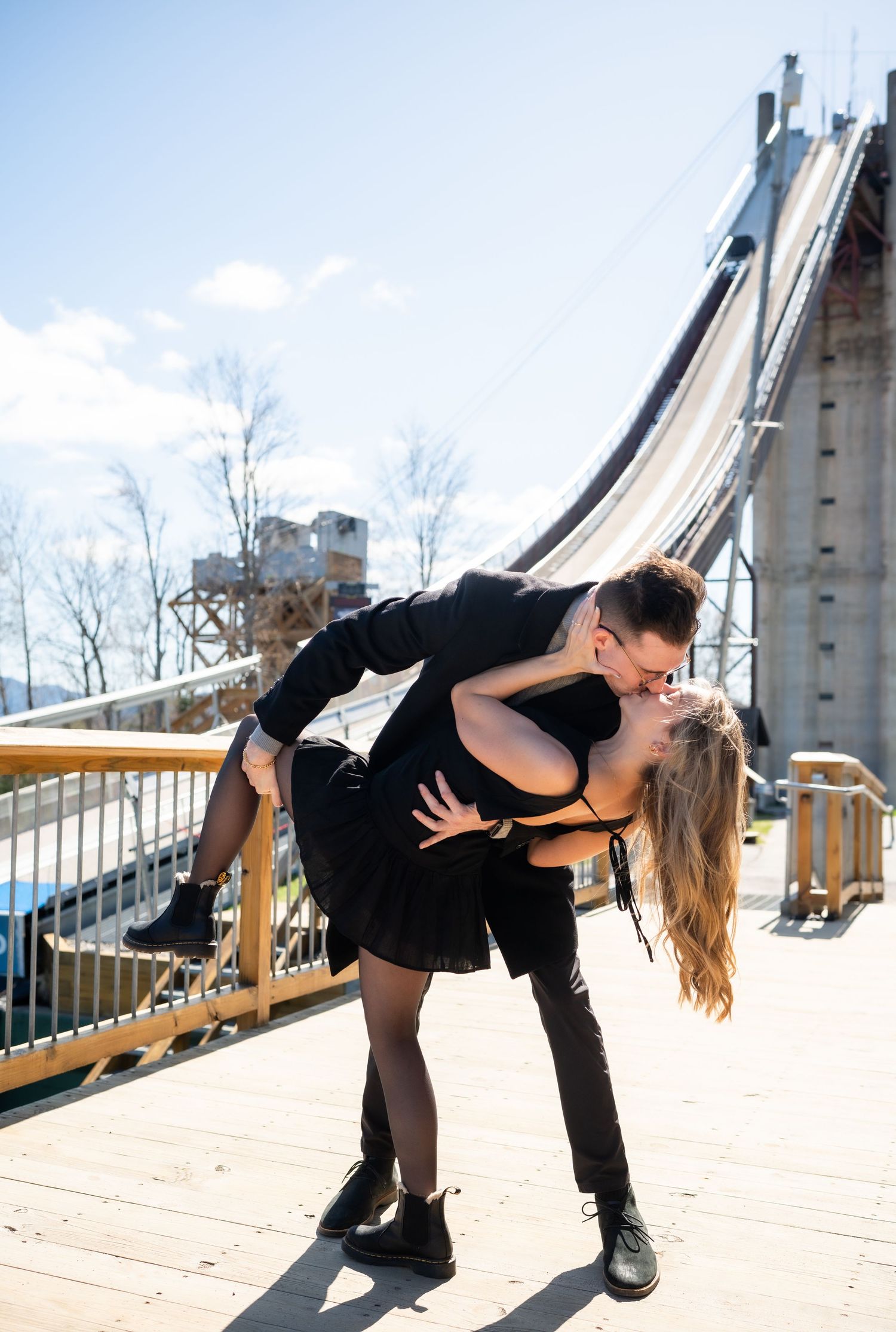 Couple in black outfits sharing a romantic dip kiss on a wooden deck with a ski jump structure behind them.