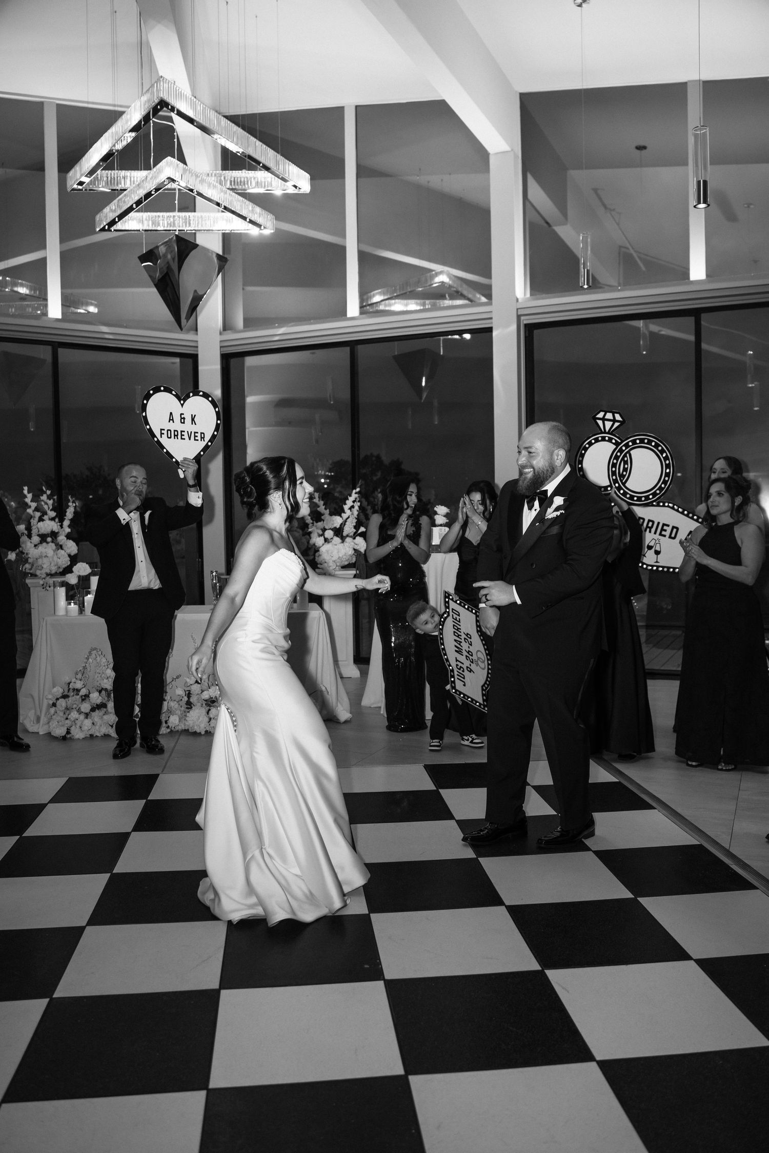Bride and groom share first dance on checkered floor at elegant wedding reception in black and white photo.