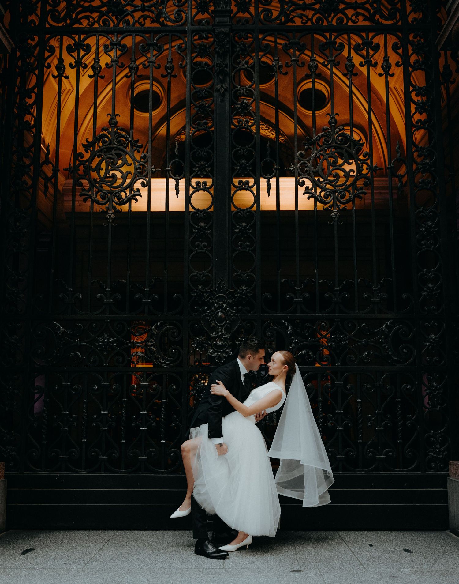 Bride and groom share a romantic moment seated before ornate black iron gates glowing warmly at dusk.