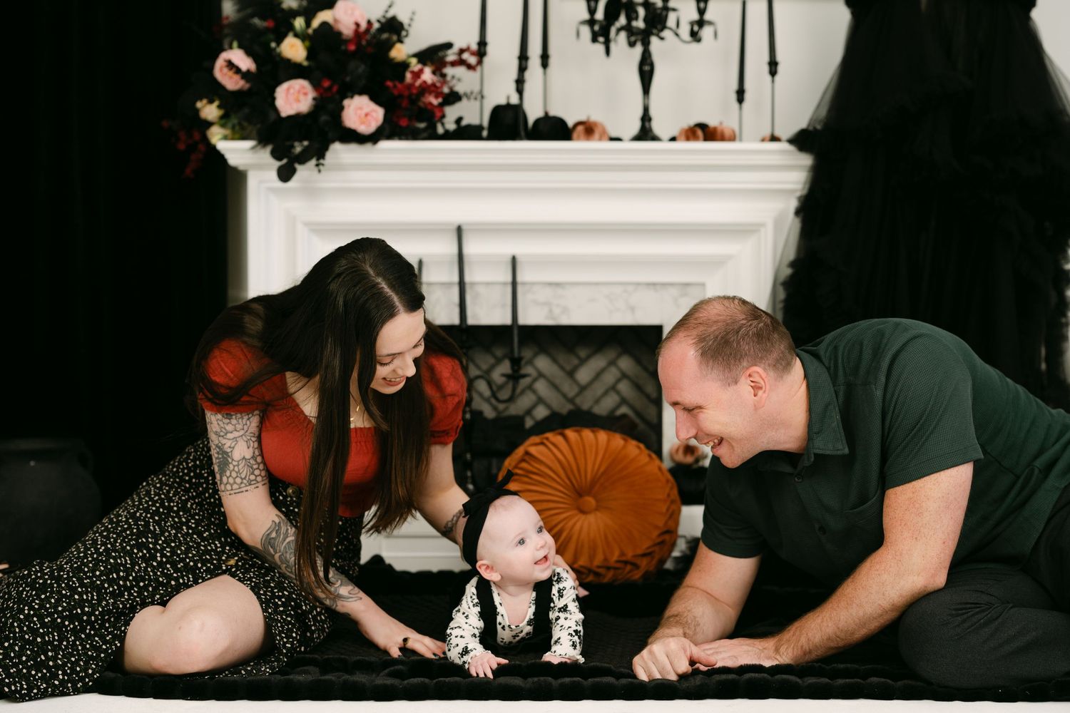 A family with a baby poses near a Halloween-decorated fireplace, parents smiling at their infant on a dark rug.