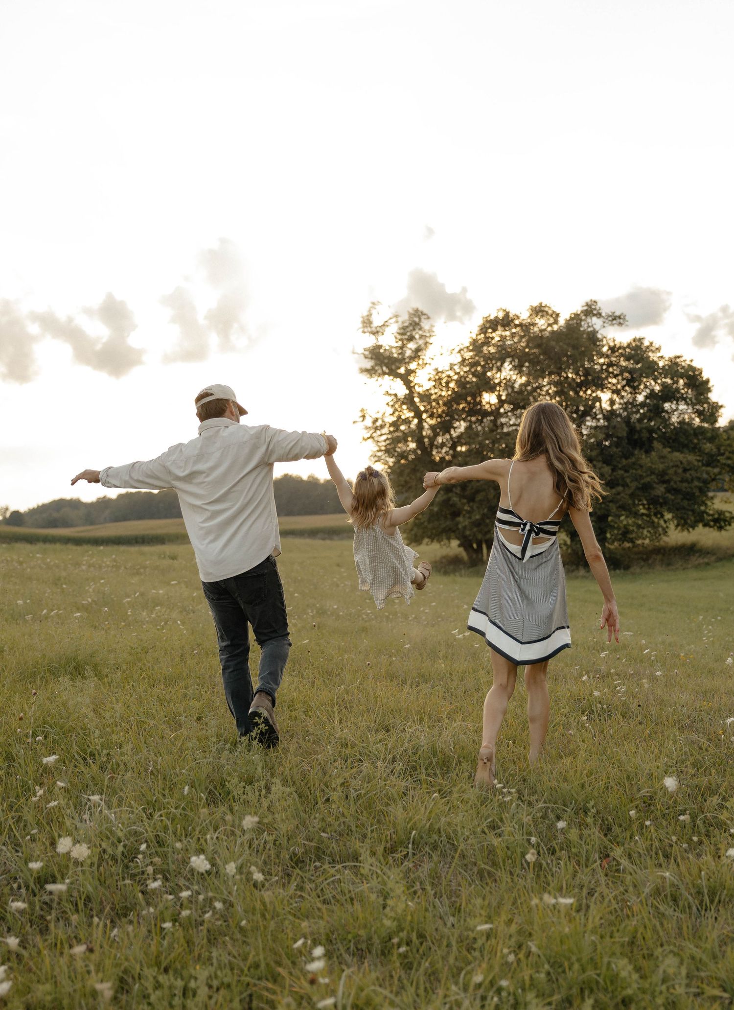 Family of three holding hands walking through a sunny green meadow, parents swinging child between them.