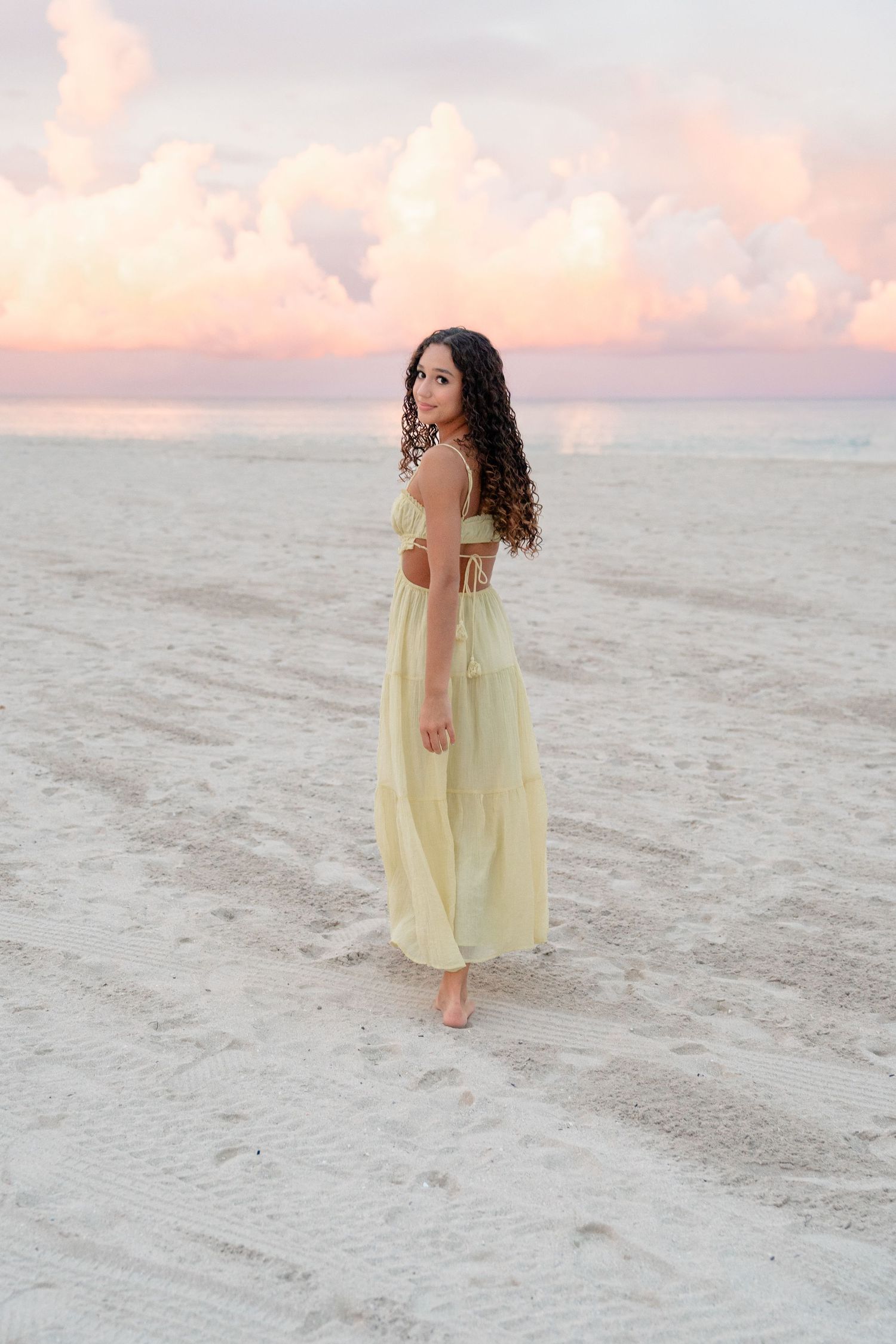 Young woman in yellow maxi dress standing barefoot on white sand beach at sunset with pink clouds.