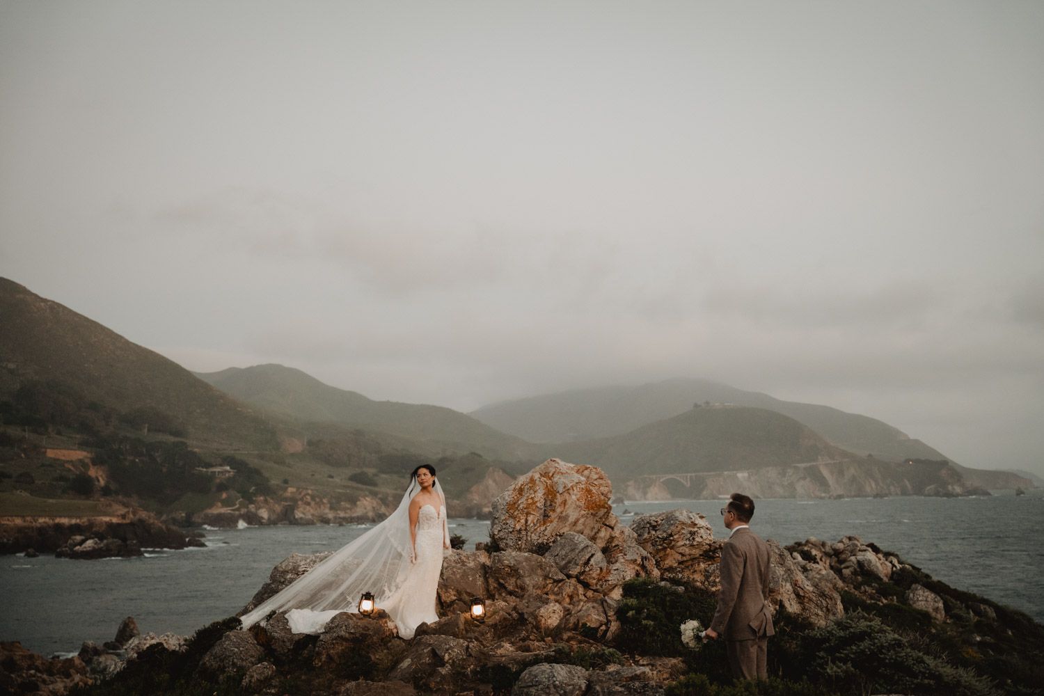 a bride in a white dress with a veil looking out in the distance on some rocks in Big Sur California with her groom looking at her