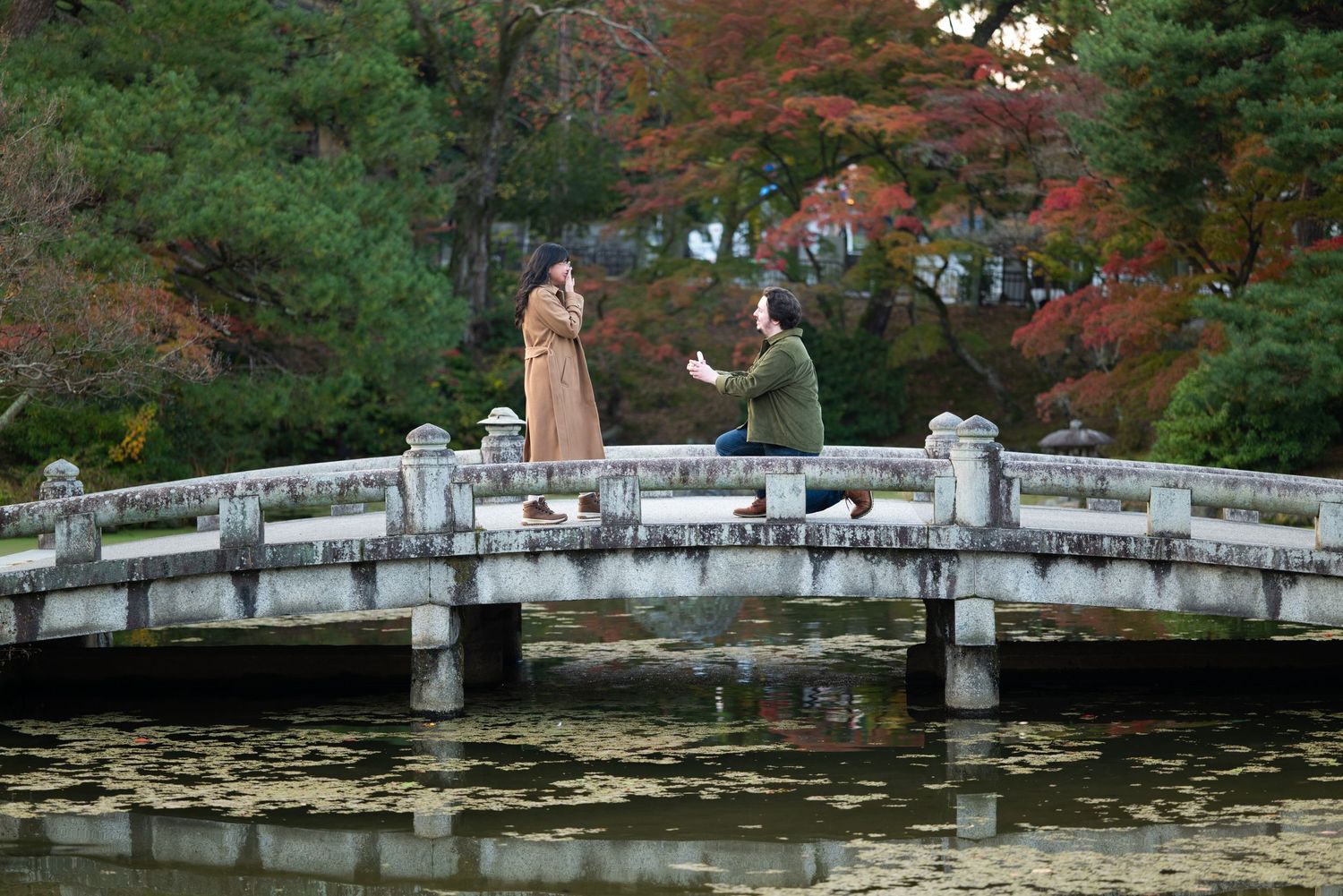 Two people stand on a stone bridge over calm water, surrounded by colorful autumn trees in a peaceful Japanese-style garden.