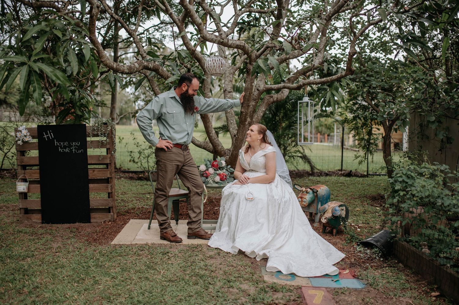 Bride in white gown and groom in blue shirt share a romantic moment under a tree at an outdoor garden wedding.