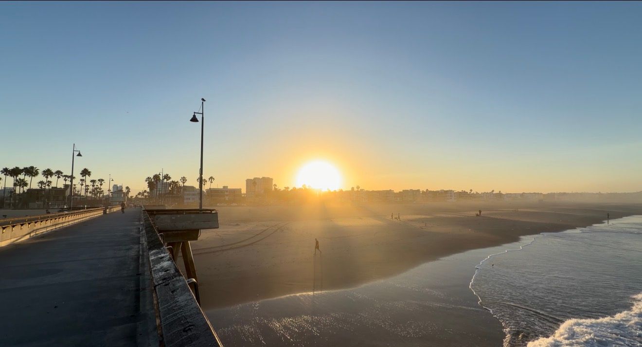 Golden sunset over a coastal pier with sandy beach and calm ocean waves in a serene seaside landscape.