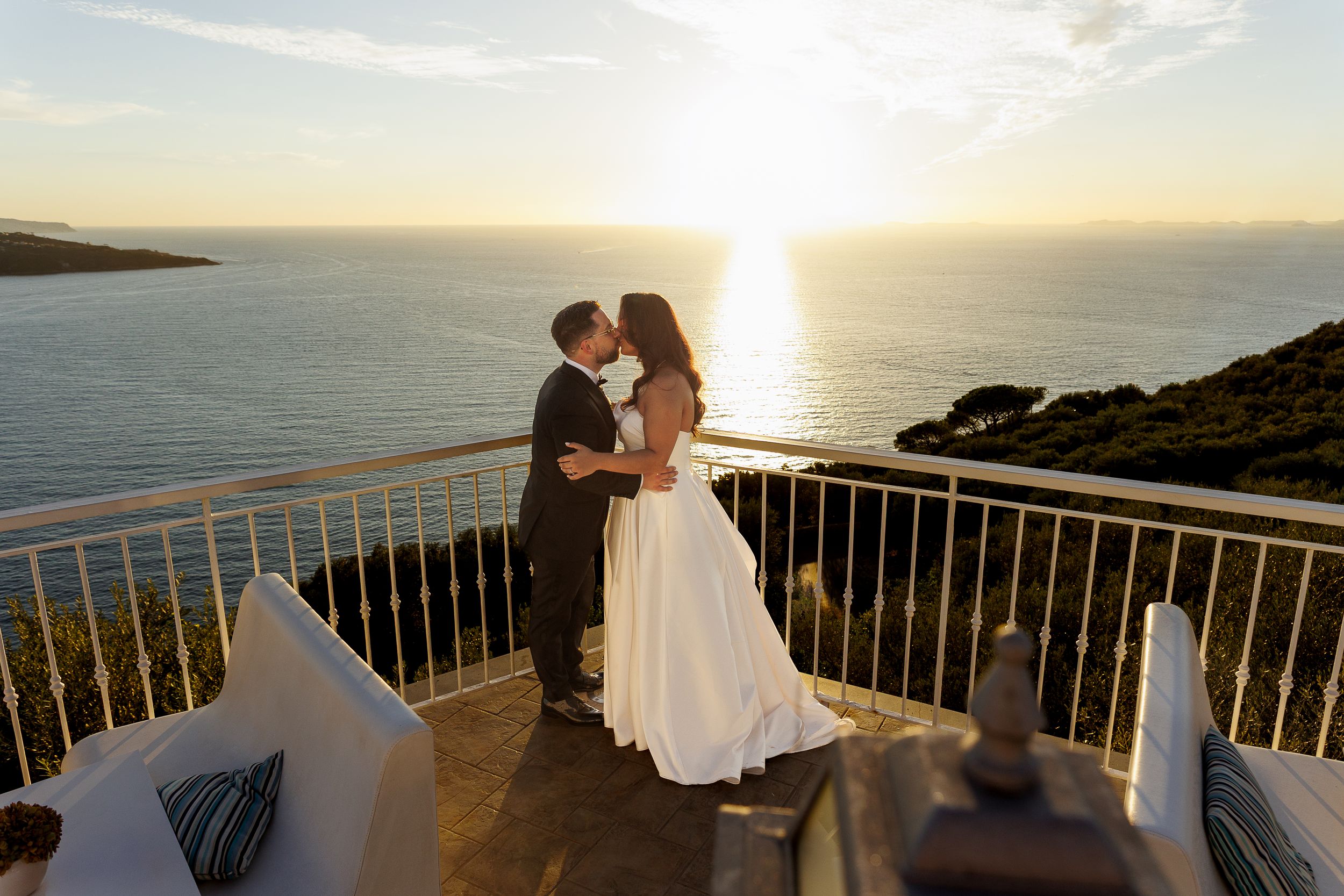 Giuliano e Barbara si baciano su una terrazza sul mare al tramonto.