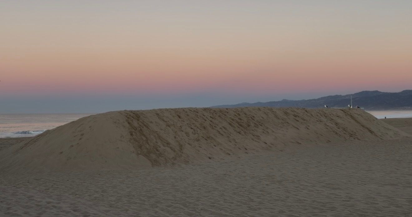 Large sand dune on a beach at sunset with a colorful pink and blue sky over the ocean horizon.